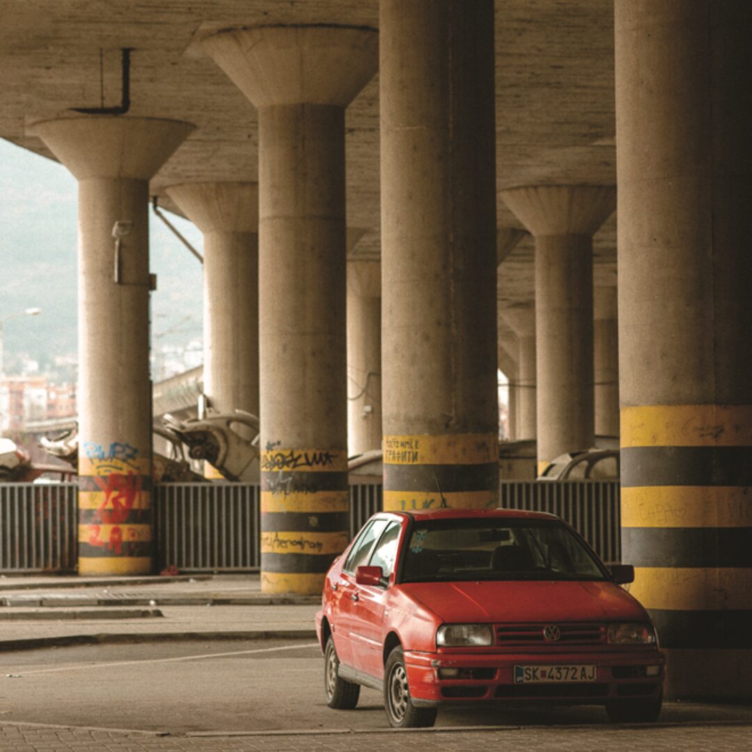 Fotografia. Czerwony pojazd zaparkowany w cieniu mostu.