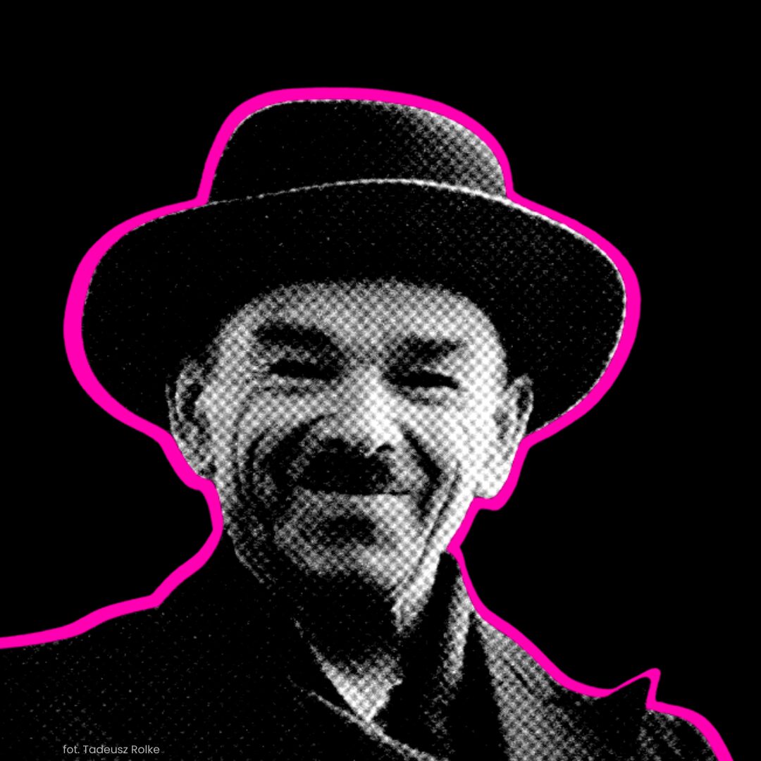 Mężczyzna w eleganckim garniturze i kapeluszu, z uśmiechem na różowym tle.