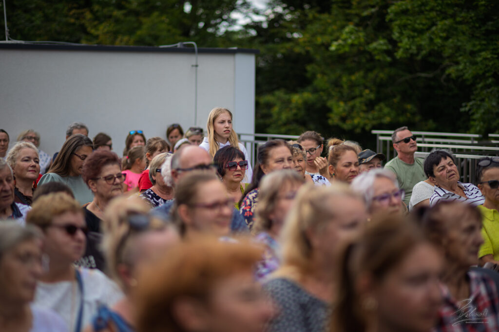 Na zdjęciu widać zróżnicowaną publiczność zgromadzoną na zewnątrz, która z uwagą obserwuje scenę.