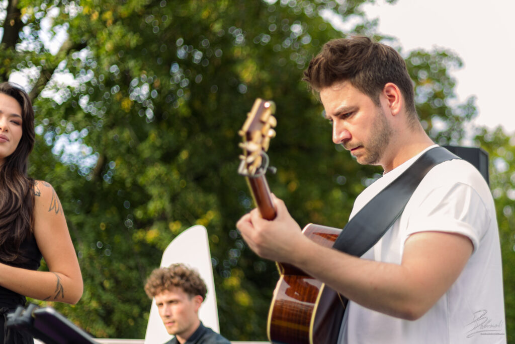 Na zdjęciu ujęcie z bliska przedstawiające gitarzystę w białym t-shircie, który z wielką precyzją i skupieniem gra na akustycznym instrumencie. W tle widać rozmytego perkusistę oraz wokalistkę, a całość dopełnia piękna, rozmyta zieleń drzew tworząca naturalną, jasną plamę w tle.