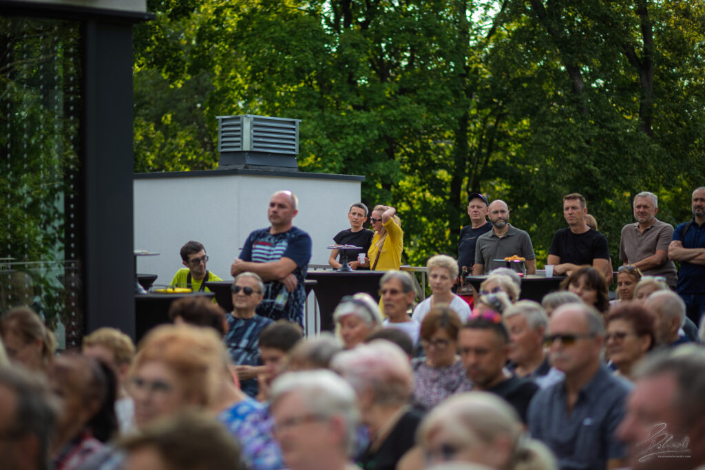 Na zdjęciu widać grupę mężczyzn stojących w głębi sceny, za plecami siedzącej publiczności. Jeden z nich w niebieskiej koszulce z charakterystycznym logo oparł się o barierkę, pozostali w czarnych i szarych koszulkach stoją swobodnie, odpoczywając i przysłuchując się muzyce w otoczeniu zieleni.