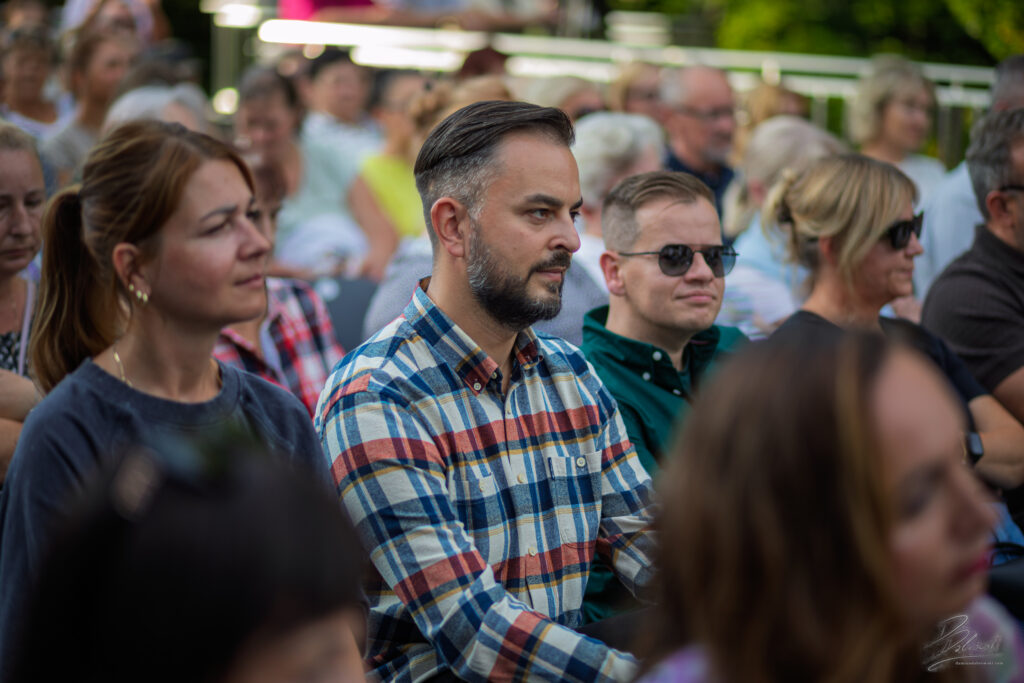 Na zdjęciu mężczyzna w kolorowej, kratowanej koszuli (niebiesko-czerwonej) siedzi na widowni i z ogromnym skupieniem patrzy na scenę. Obok niego siedzi kobieta w ciemnej bluzie, a za nimi pozostali słuchacze, którzy z uwagą śledzą przebieg koncertu.