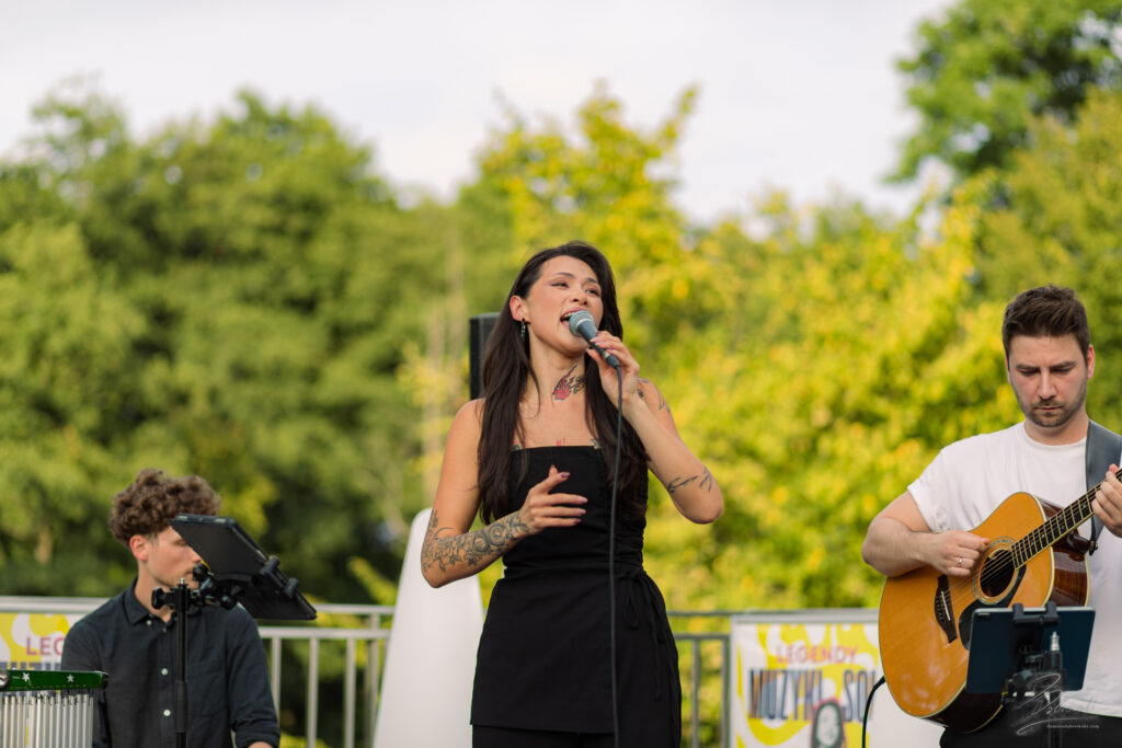 Na zdjęciu wokalistka trzyma mikrofon, ma zamknięte oczy i wyraźnie wzruszona śpiewa, kładąc dłoń na sercu. Obok niej stoi gitarzysta, a za nimi gra na instrumencie perkusyjnym młody mężczyzna. Na twarzy wokalistki widać ogromne emocje i pasję do muzyki.