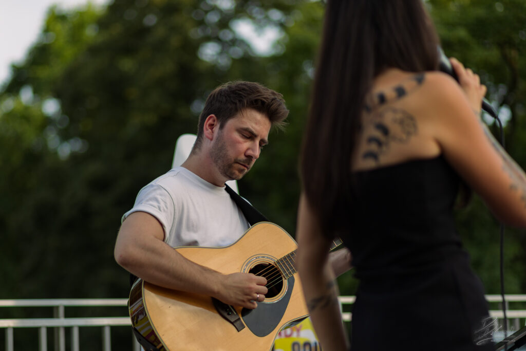 Na zdjęciu zbliżenie na gitarzystę w białej koszulce, który z pochyloną głową gra na akustycznej gitarze. W tle widoczna jest rozmyta sylwetka wokalistki z tatuażem, a naturalne światło i zieleń drzew tworzą przyjemny, spokojny klimat koncertu.