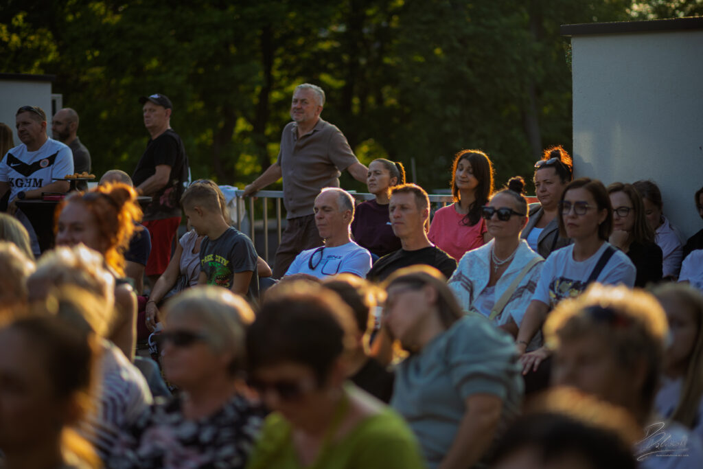 Na zdjęciu widać publiczność zgromadzoną na widowni w ciepłym, wieczornym świetle. Twarze słuchaczy są rozświetlone złotym blaskiem zachodzącego słońca, a wśród nich wyróżnia się kobieta o rudych włosach, która uśmiecha się, oklaskując artystów na scenie.