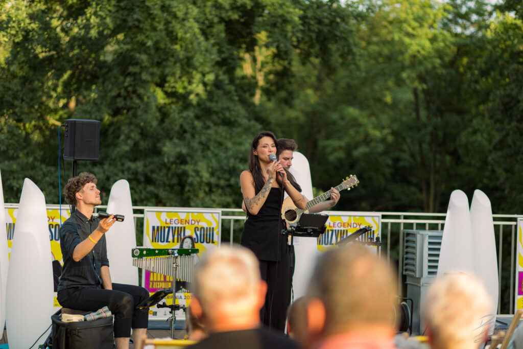 Na zdjęciu ujęcie zza pleców publiczności, która obserwuje występ zespołu (wokalistka, perkusista z pałeczkami w dłoniach i gitarzysta). Widać wyraźnie, jak koncert zgromadził wielu słuchaczy, a scena z białymi kolumnami idealnie komponuje się z zielenią.
