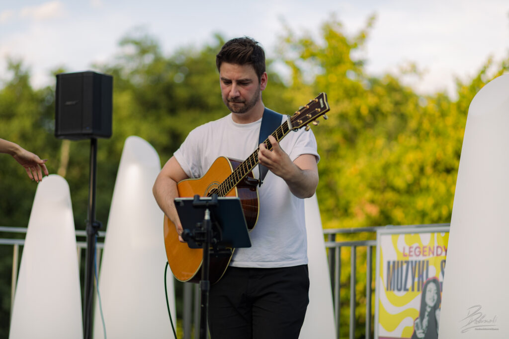 Na zdjęciu gitarzysta w białym t-shircie, ubrany w czarne spodnie, gra na akustycznym instrumencie, patrząc przed siebie. Za nim znajduje się statyw z nutami oraz białe, dekoracyjne płyty, które tworzą niepowtarzalną scenografię tego plenerowego wydarzenia.