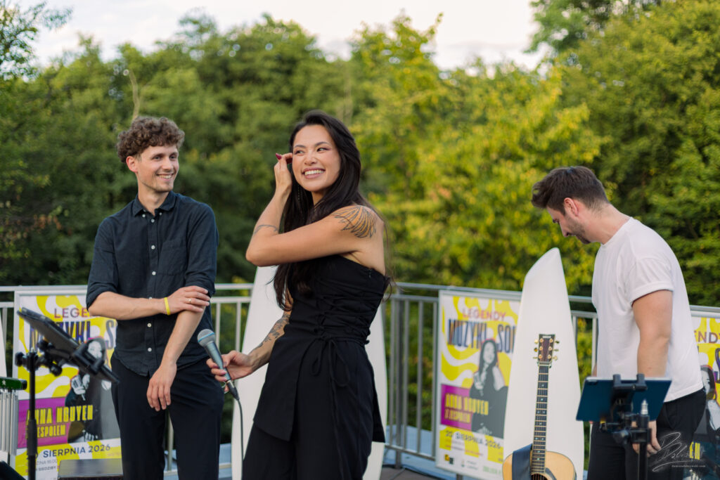 Na zdjęciu wokalistka, perkusista i gitarzysta stoją na scenie, uśmiechając się do siebie po zakończonym występie. Na ich twarzach widać radość i satysfakcję ze wspólnego grania, a tło stanowią kolorowe plakaty koncertowe.