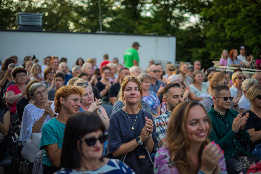 Na zdjęciu szeroki plan przedstawiający publiczność, która nagradza artystów gromkimi brawami. Widać uśmiechnięte twarze i klaszczące dłonie, a muzycy na scenie dziękują za wspaniałe przyjęcie, co wieńczy to wyjątkowe, letnie spotkanie.
