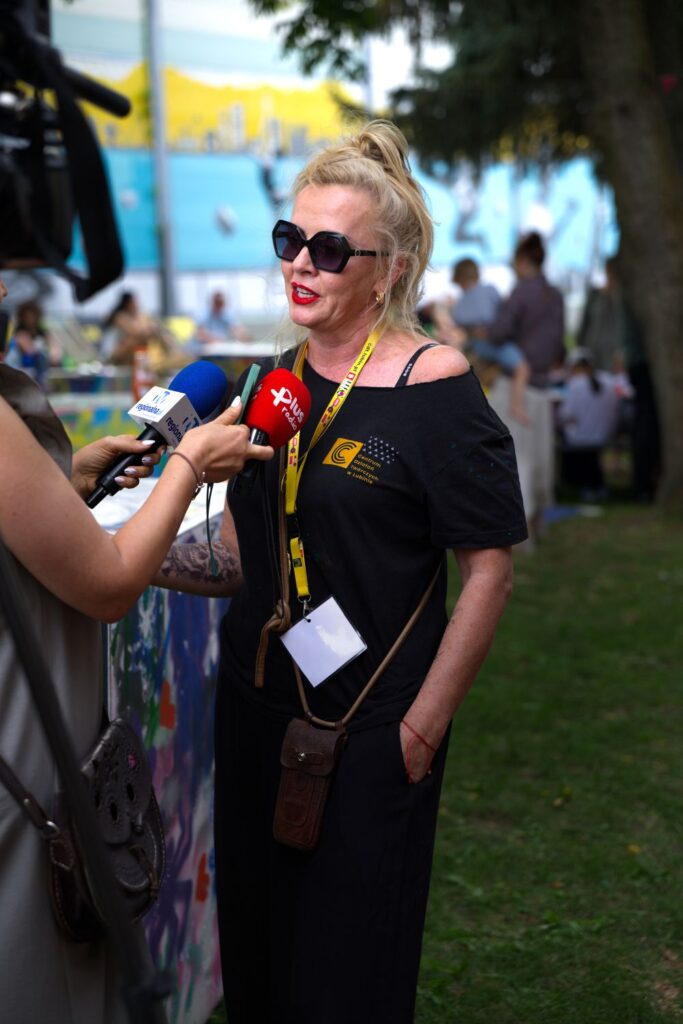 Na fotografii kobieta w czarnej koszulce z logo CDT i okularach przeciwsłonecznych - Dyrektor CDT Anna Wiśniewska - udziela wywiadu, odpowiadając na pytania dziennikarzy trzymających mikrofony.