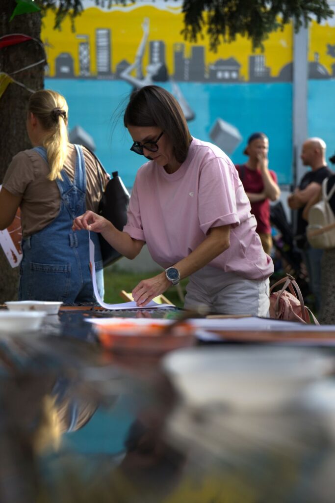 Na fotografii kobieta w różowym t-shircie i okularach pochyla się nad stołem, podziwiając swoją pracę plastyczną.