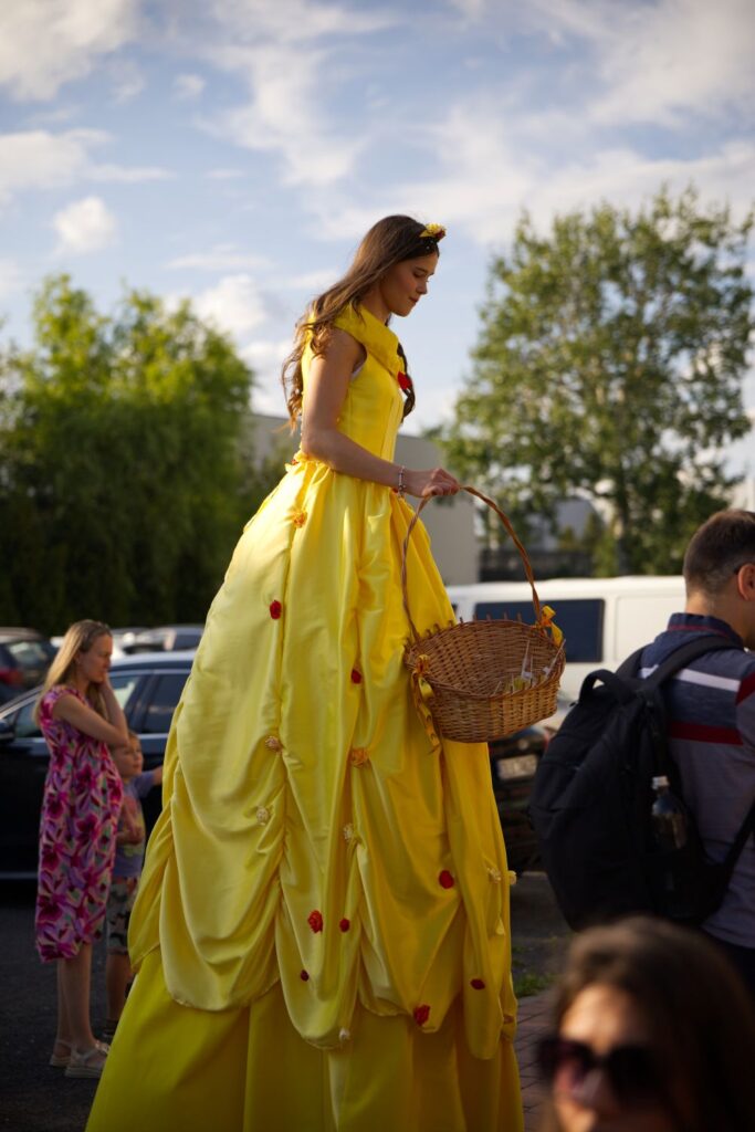Na fotografii kobieta na szczudłach ubrana w wielką, żółtą suknię z różami - strój księżniczki - przechadza się wśród uczestników pikniku.