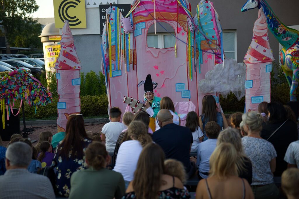 Na fotografii z perspektywy widowni widać aktorów występujących na kolorowej scenie, a przed nimi siedzą dzieci na kolorowych poduszkach.