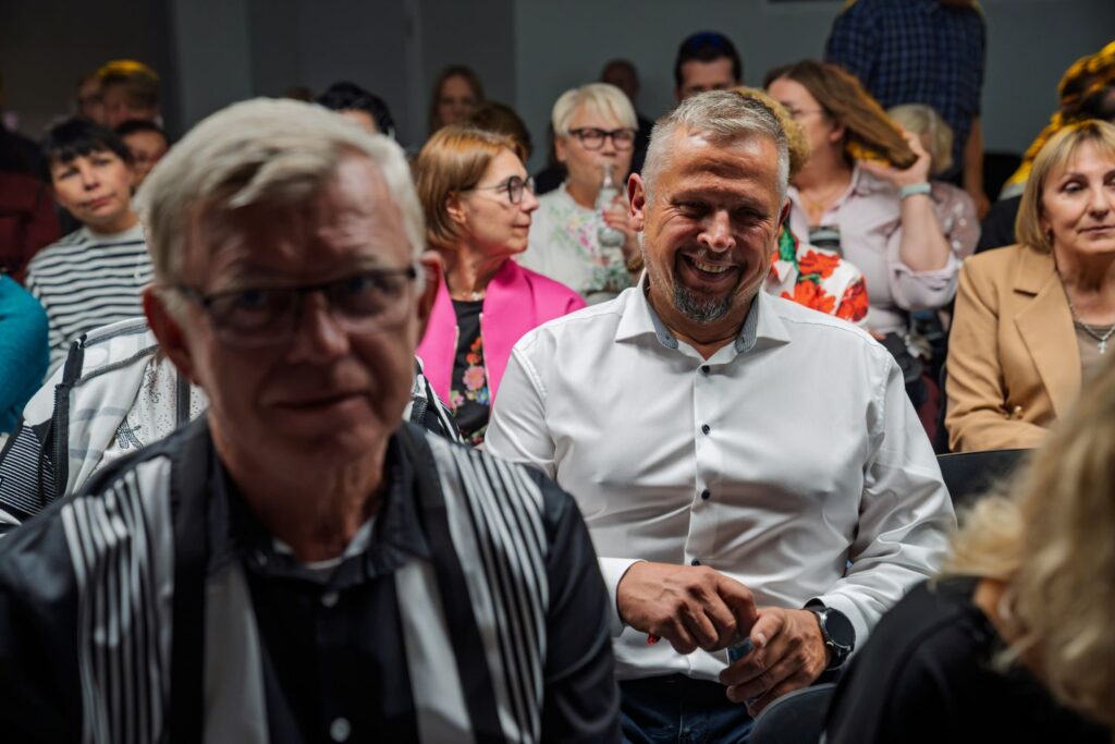 Mężczyzna w białej koszuli z uśmiechem na twarzy wśród publiczności.
