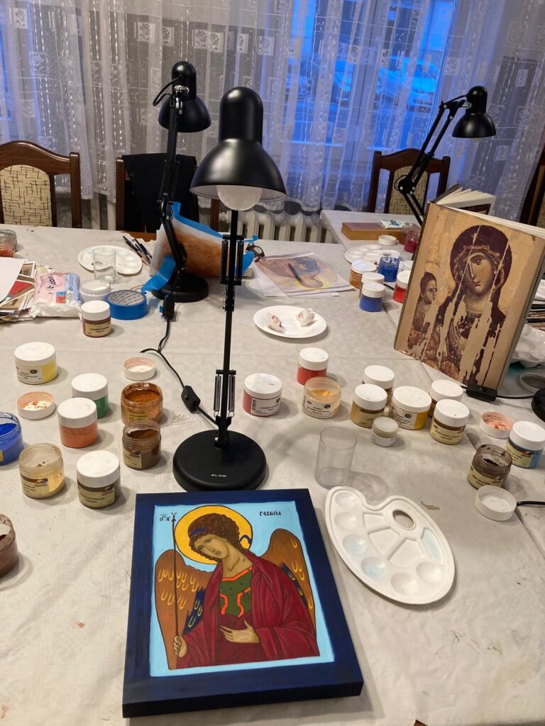 Na zdjęciu na stole leży ikona anioła w czerwonej szacie na niebieskim tle, a wokół niej rozłożone są farby, palety i lampka biurkowa.