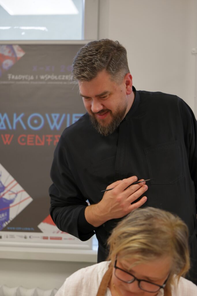 Uśmiechnięty mężczyzna z pędzlem w ręku na tle plakatu festiwalu.
