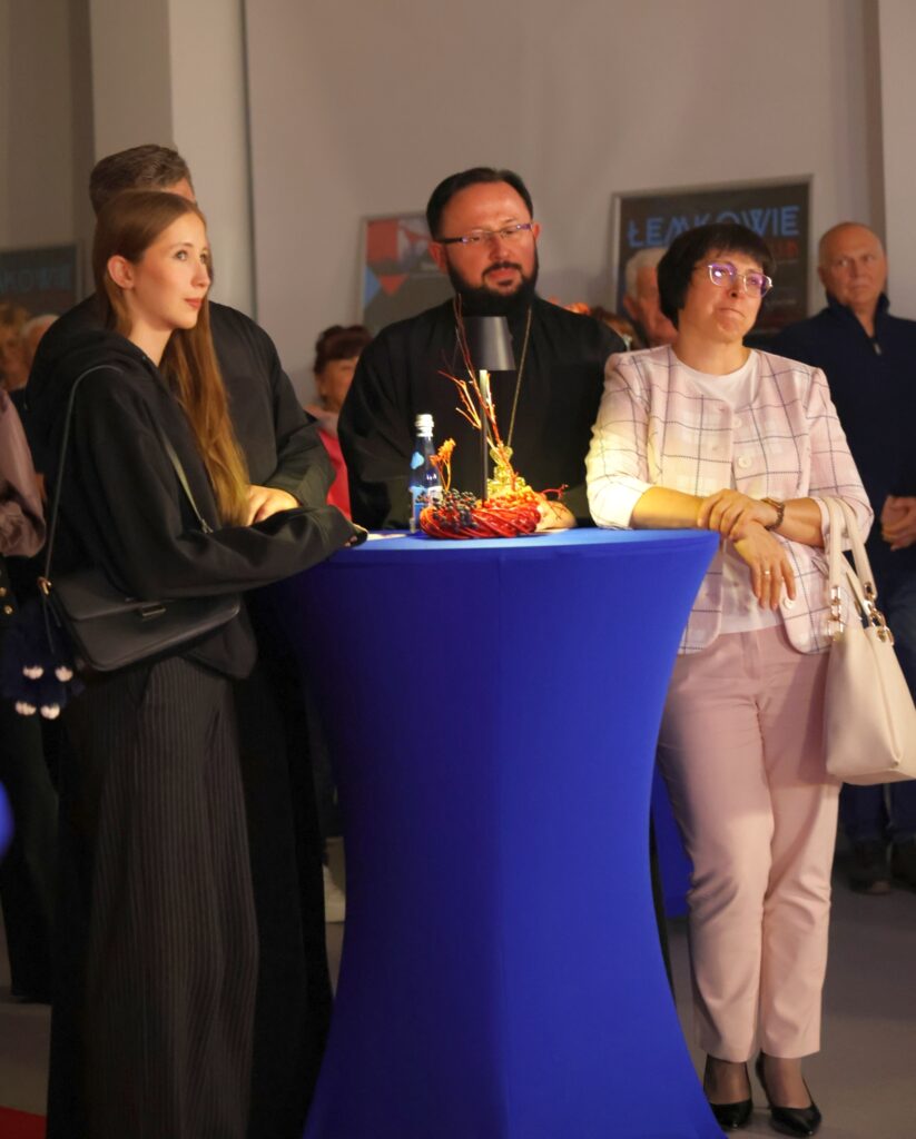 Zdjęcie przedstawia ludzi zgromadzonych przy niebieskim stoliku, a wśród nich mężczyznę w sutannie i z brodą.