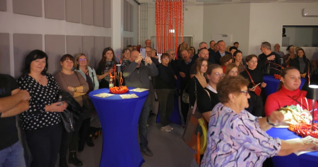 Na fotografii widać licznych uczestników wernisażu stojących w holu, z osobą w środku nagrywającą wydarzenie smartfonem.