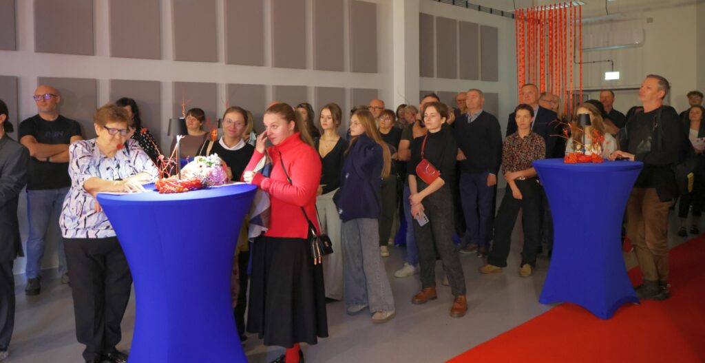 Zdjęcie przedstawia dużą grupę osób stojących w sali przy niebieskich stolikach, z uwagą słuchających wystąpień.