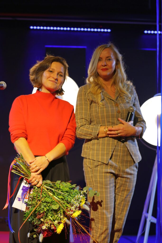 Na fotografii widać dwie kobiety na scenie, jedną w czerwonym swetrze trzymającą kwiaty i drugą w kraciastym garniturze z mikrofonem.