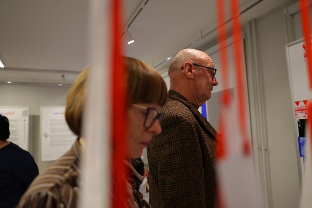 Na fotografii widać starszego mężczyznę w okularach oglądającego ekspozycję, sfotografowanego przez pionowe, czerwone wstążki.