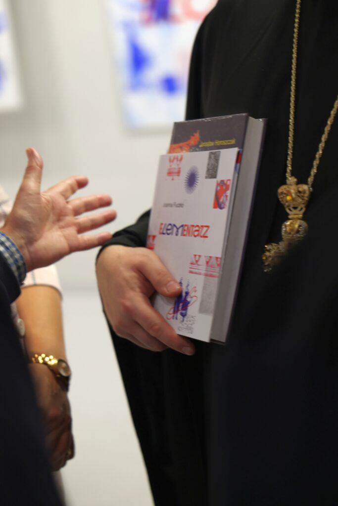 Na fotografii widać osobę w czarnym stroju trzymającą publikację „Elementarz”, obok której znajduje się dłoń drugiej osoby gestykulującej w rozmowie.