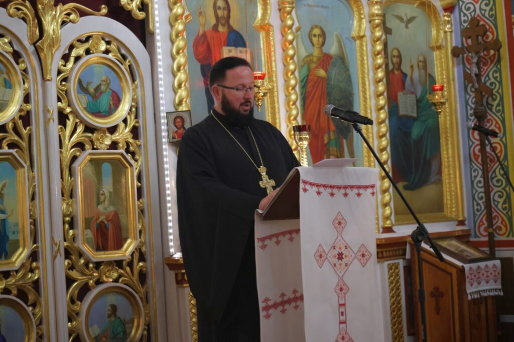 Duchowny w okularach (ks. Bogdan Repeła) stoi przy mównicy w cerkwi, zapowiadając zespół.