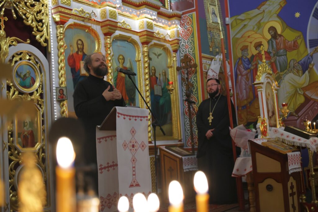 Członek chóru z brodą gestykuluje przy mikrofonie, obok stoi duchowny. Na pierwszym planie widać płomienie świec.
