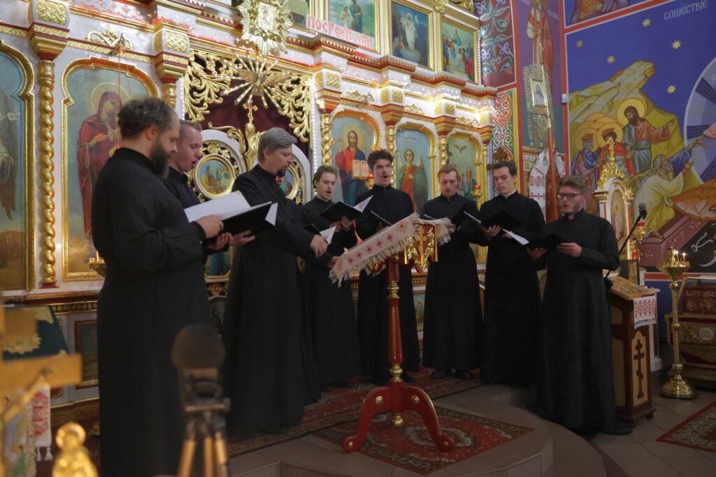 Męski chór w czarnych szatach śpiewa z nut, stojąc przed ikonostasem.