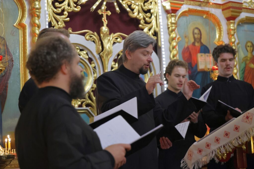 Dyrygent w czarnej szacie prowadzi śpiew męskiego chóru w cerkwi.