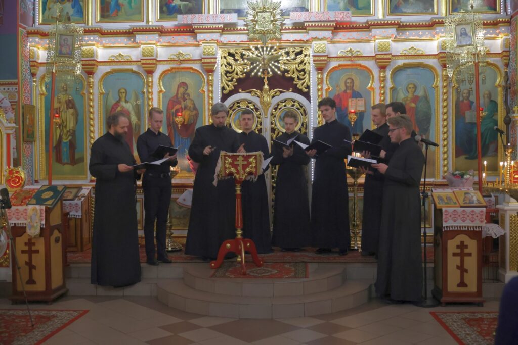 Chór męski w czarnych strojach śpiewa, stojąc wokół pulpitu z krzyżem.