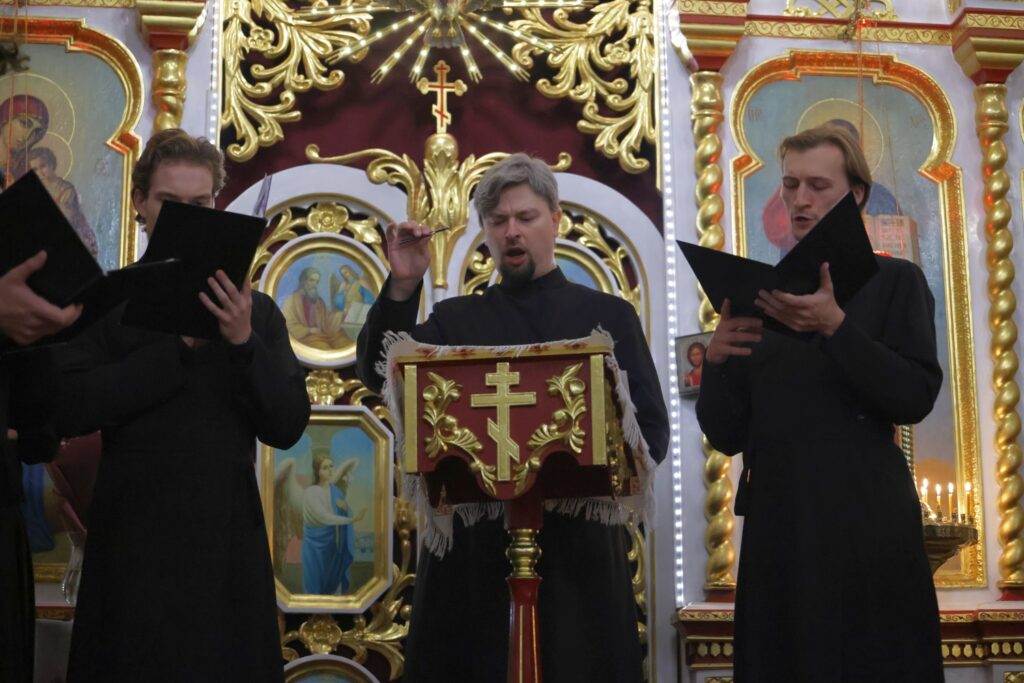 Dyrygent z batutą prowadzi chór, obok śpiewacy trzymają czarne nuty.