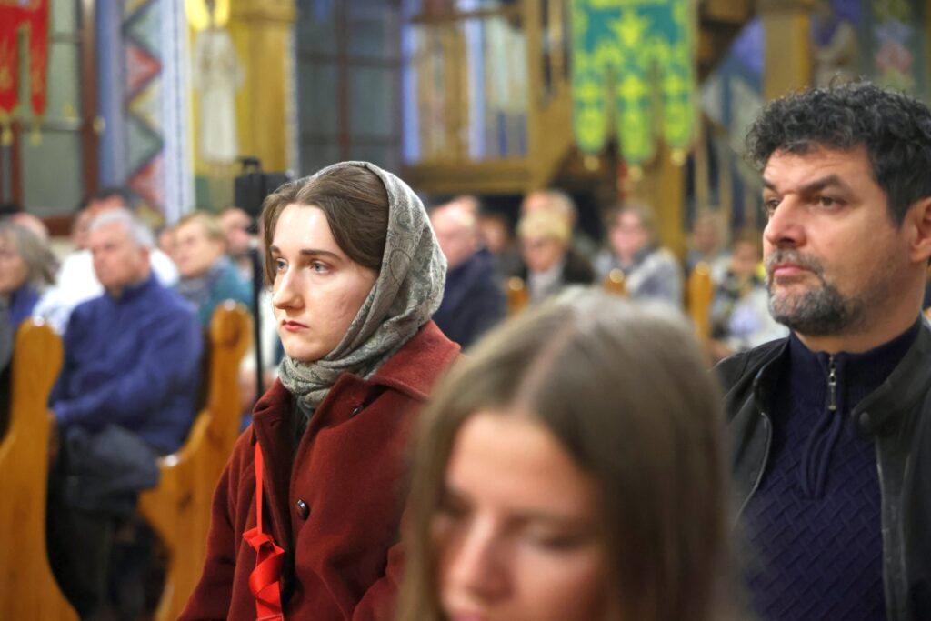 Portret kobiety w chuście i czerwonym płaszczu słuchającej koncertu w cerkwi.