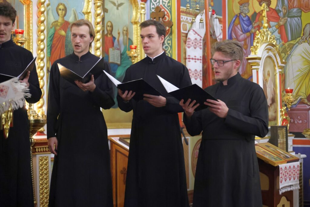 Trzech młodych śpiewaków w czerni z otwartymi nutami na tle złotych ikon.