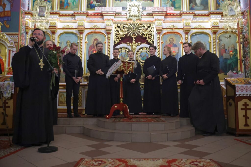 Duchowny z mikrofonem i bukietem czerwonych róż zwraca się do słuchaczy. Tuż za nim stoi zespół.