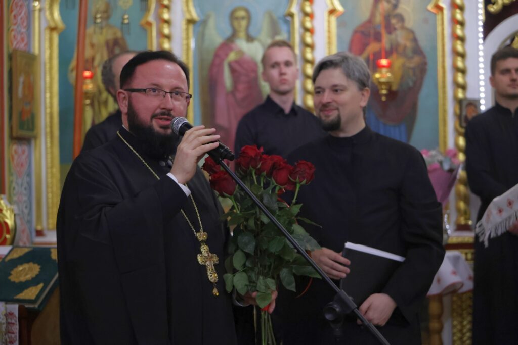 Duchowny z mikrofonem i bukietem czerwonych róż zwraca się do słuchaczy. Tuż za nim stoi zespół.