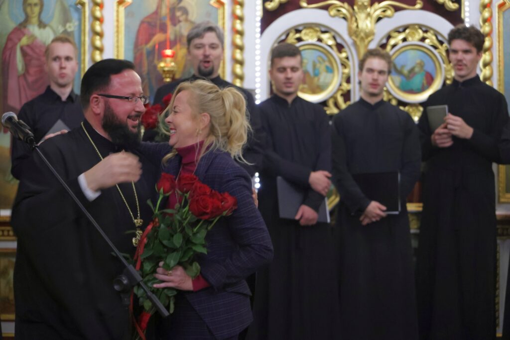Duchowny wręcza bukiet czerwonych róż kobiecie w granatowym garniturze.