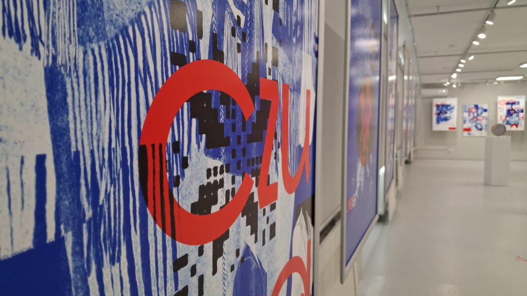 Zdjęcie przedstawia detal plakatu z dużymi, czerwonymi literami na niebieskim tle, ukazany w perspektywie ciągnącego się korytarza wystawowego.