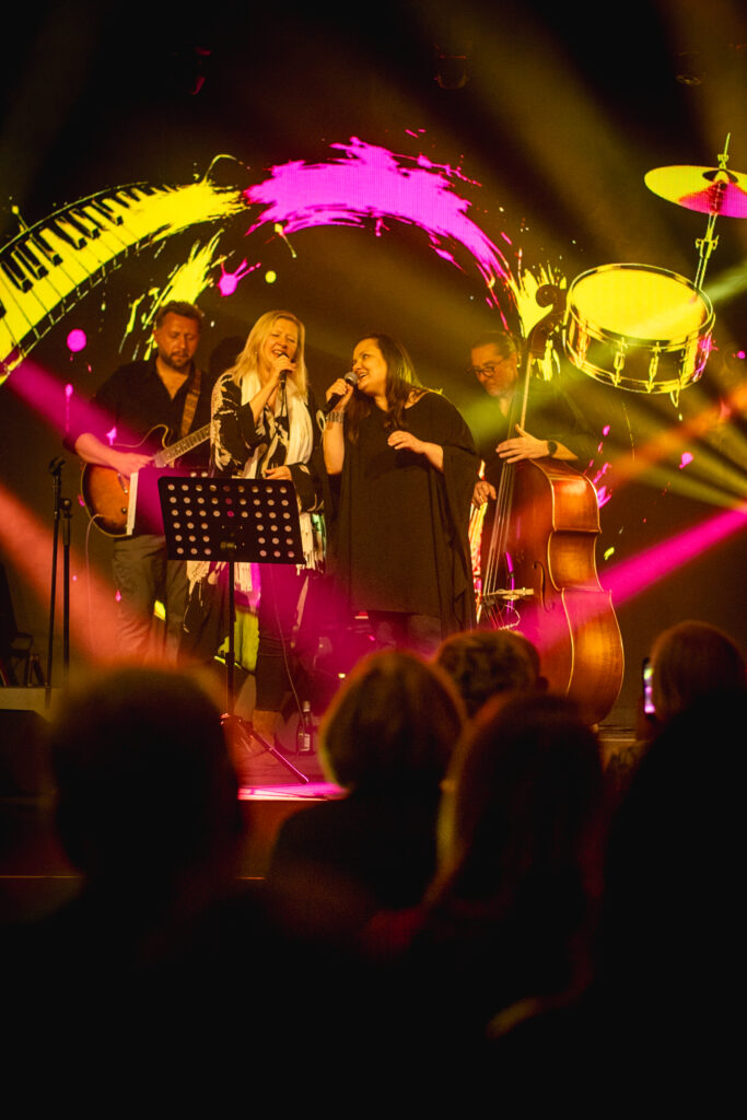 Na zdjęciu zespół jazzowy występuje na scenie, a wokalistki śpiewają do mikrofonów w otoczeniu dynamicznych świateł i sylwetek publiczności.