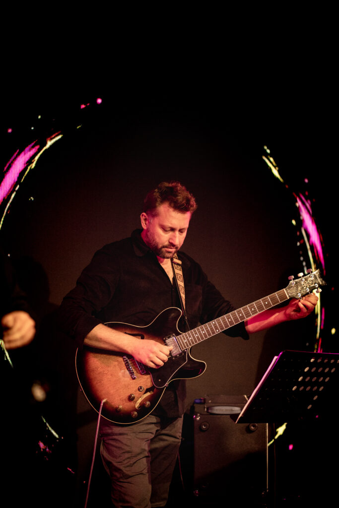 Na zdjęciu gitarzysta w czarnej koszuli gra na gitarze, oświetlony scenicznym światłem.