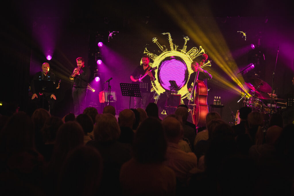 Na zdjęciu zespół jazzowy występuje na scenie na tle dużego, świecącego konturu z motywem instrumentów.