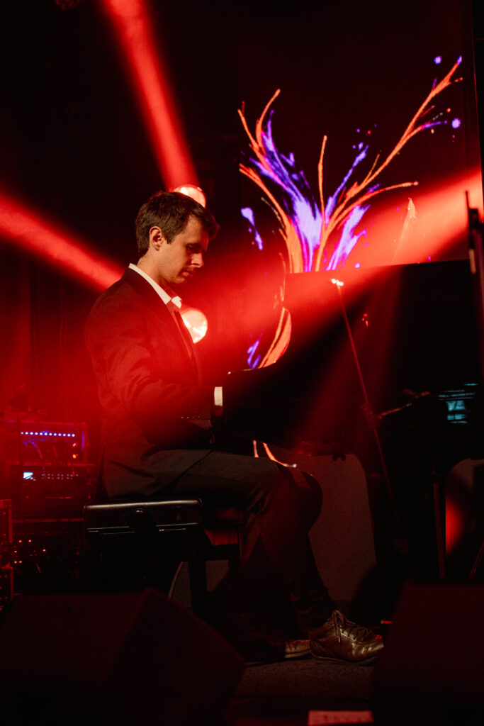 Na zdjęciu pianista w garniturze gra na fortepianie, oświetlony intensywnym czerwonym światłem.