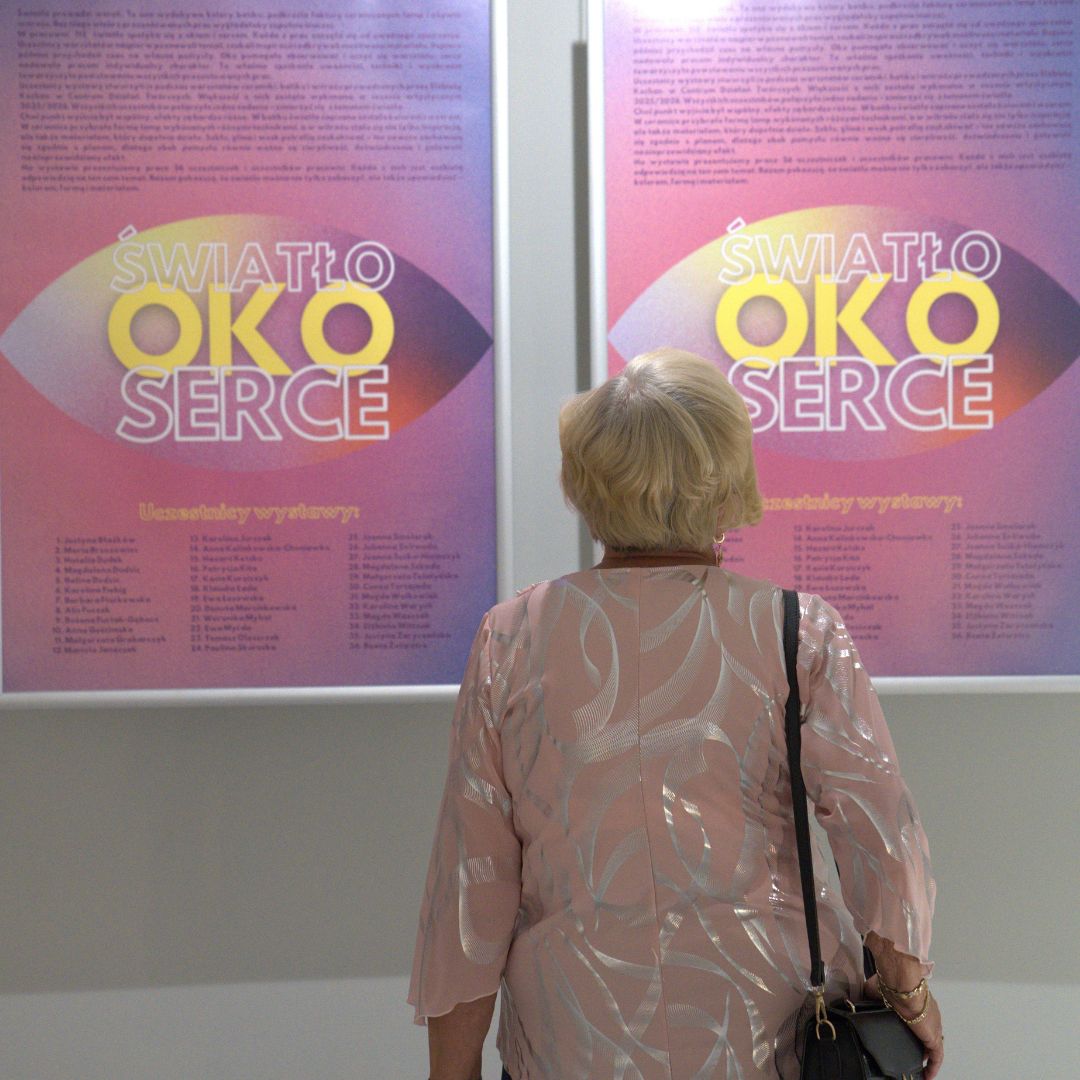 Starsza kobieta w różowej bluzce ogląda plakaty wystawy „Światło. Oko. Serce." z listą uczestników